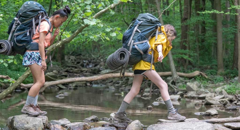 Two people wearing backpacks cross a creek over stones